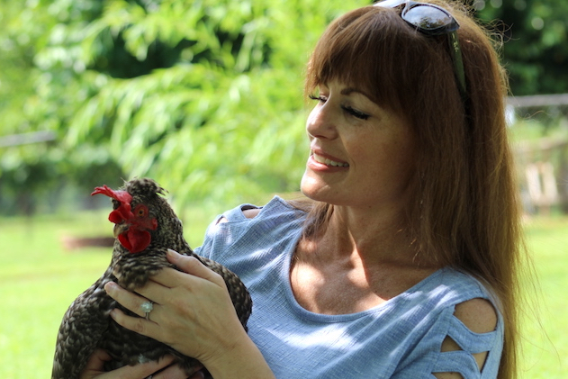 woman with long dark hair in blue shirt holding a black and white speckled chicken outside