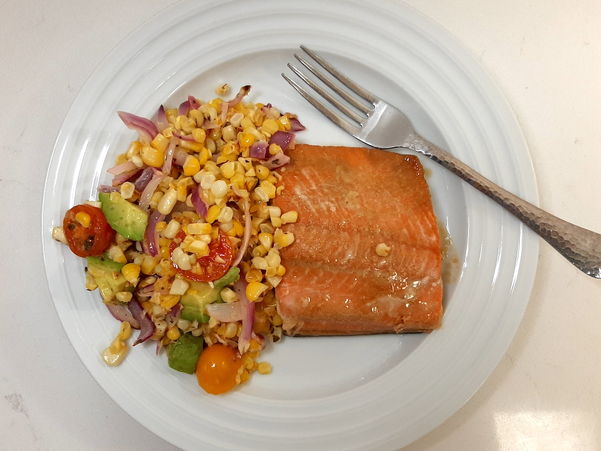 cooked salmon with corn kernels, sliced red onion, cherry tomatoes, and diced avocado on a white plate on white background