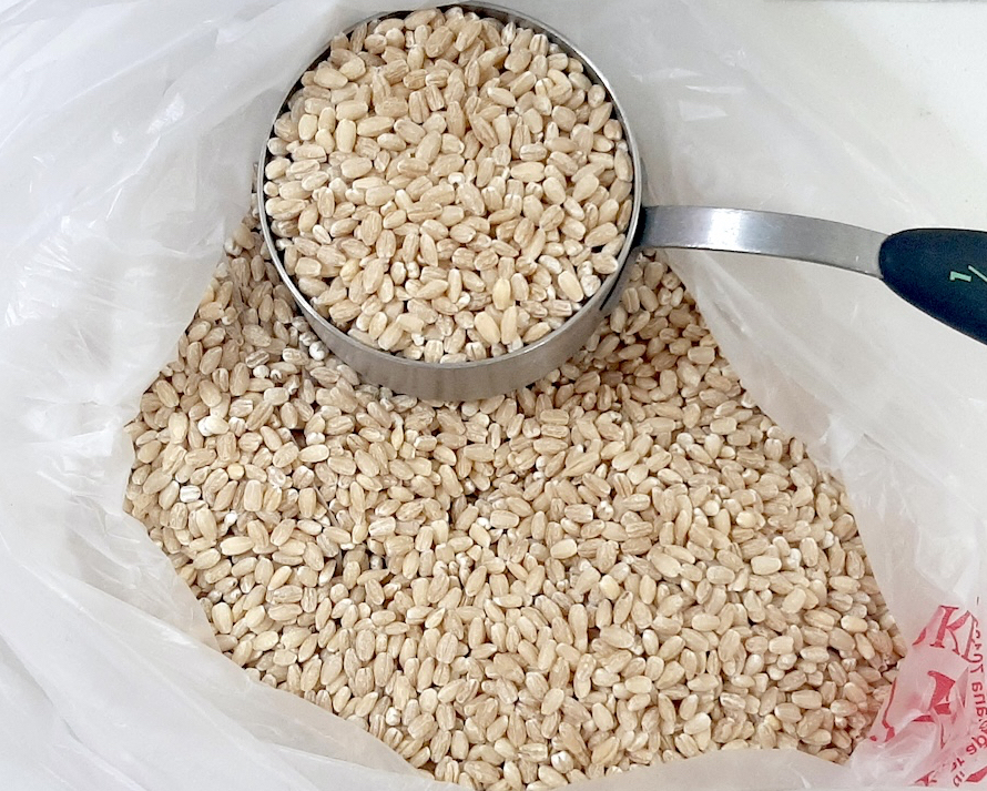 some type of beige grain in a measuring cup on top of more grain sitting on a clear plastic bag on a white background