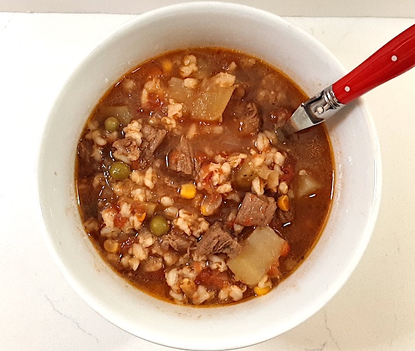 cooked beef and vegetable soup in a white bowl with a red spoon on a white background